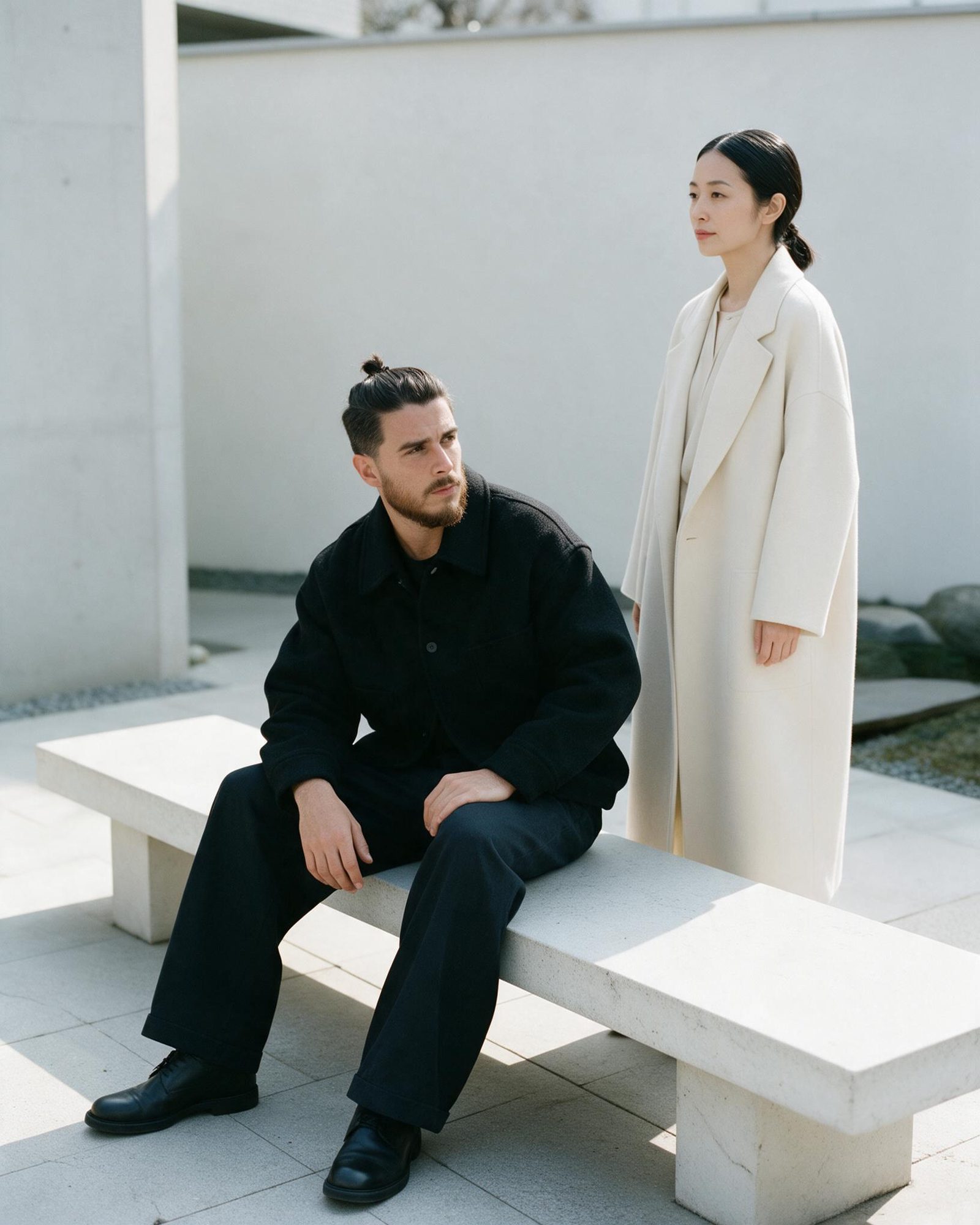 Xavier Roca seated on a stone bench beside a model in a cream coat