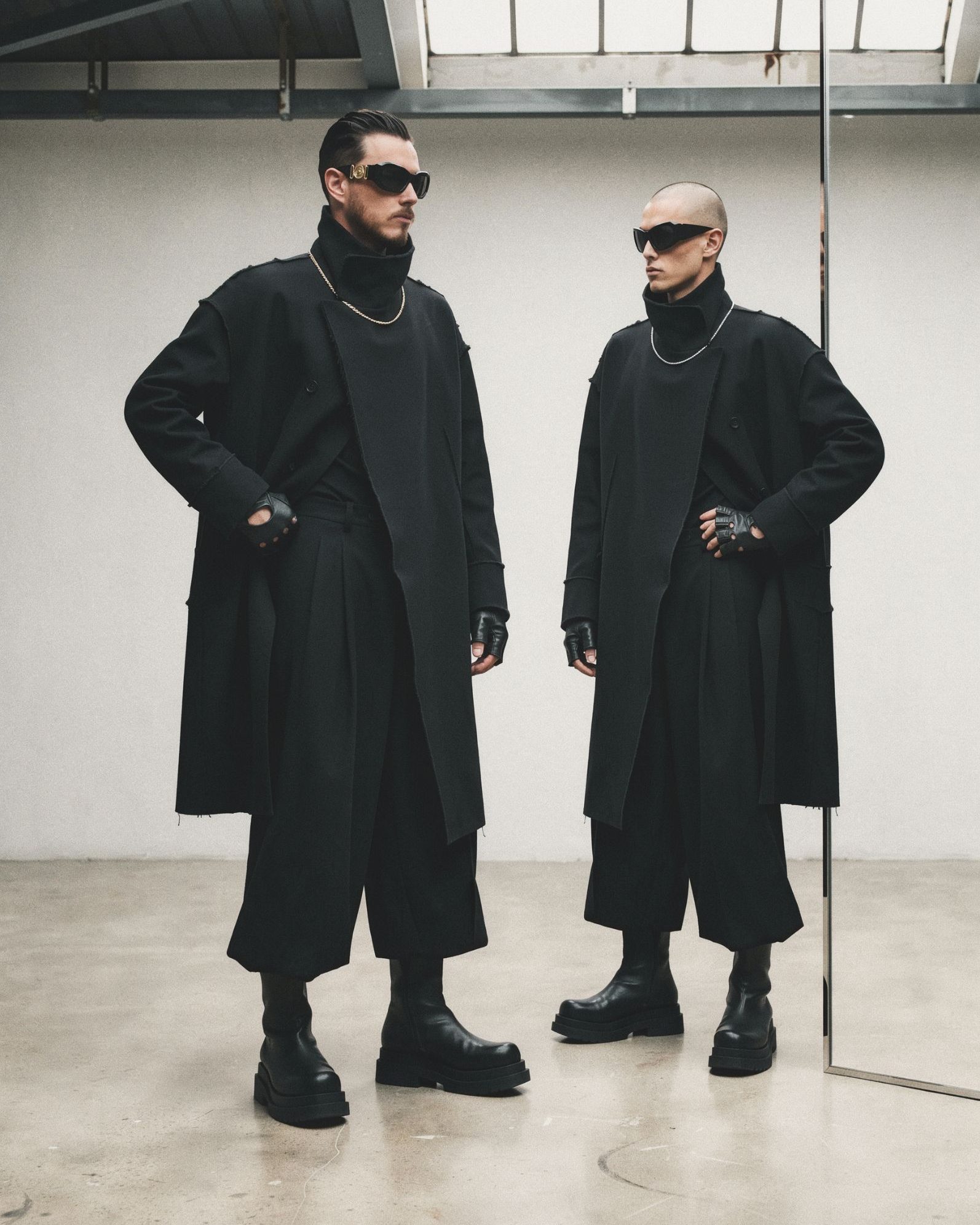 Xavier Roca and a second model in matching long black coats, wide pleated trousers, fingerless gloves and platform boots in a white studio