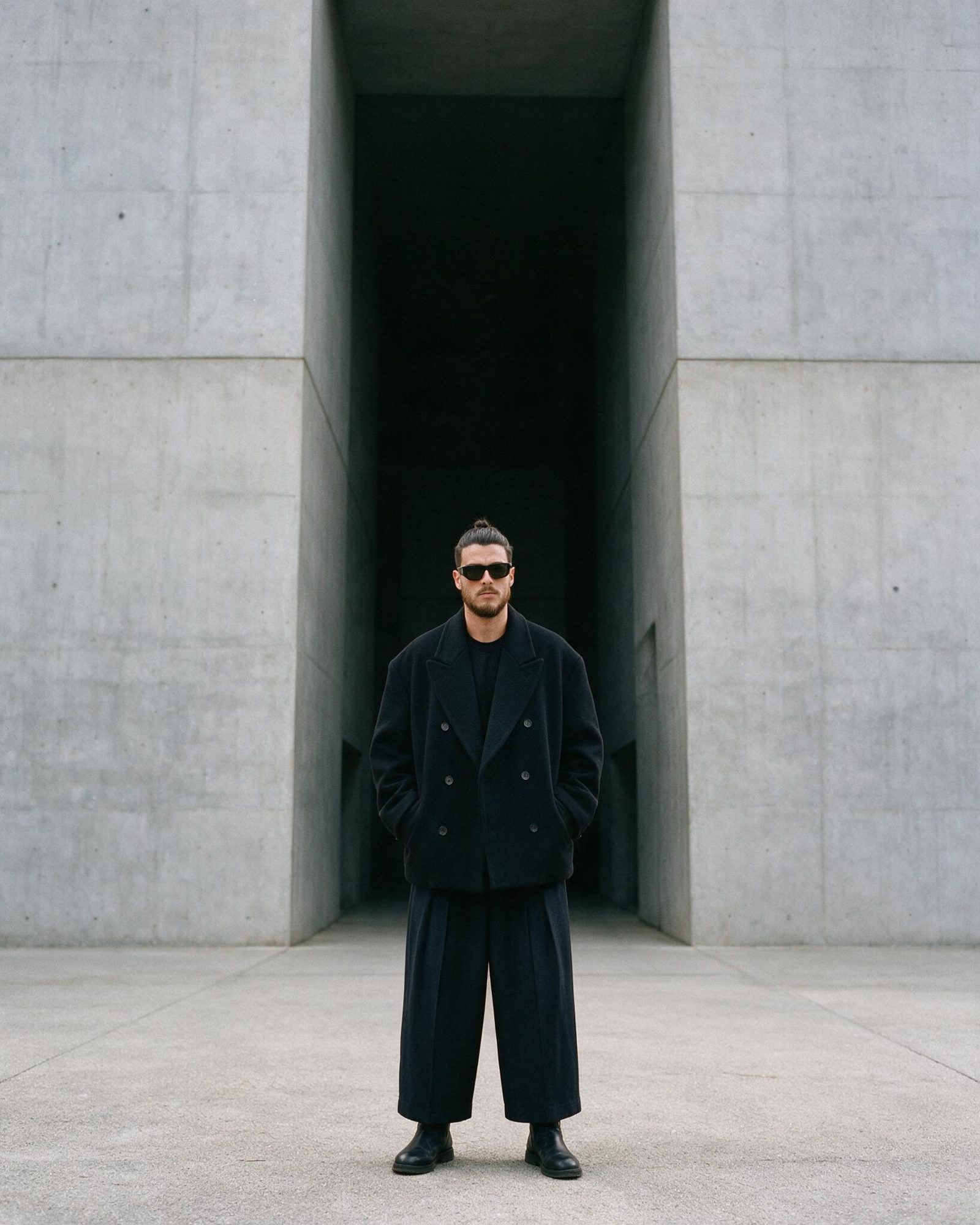 Xavier Roca in a black double-breasted coat and wide trousers, in front of a concrete passage